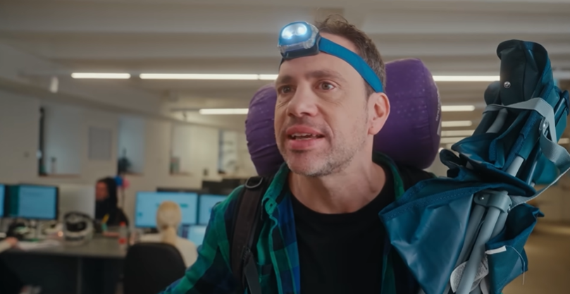 Office worker dressed as camper with headlamp and backpack in SysAid AI agents commercial