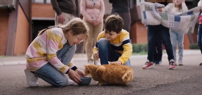 Two children kneeling on pavement feeding a small brown dog outside apartment building