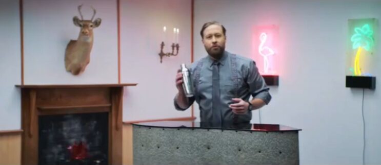 Man holding a cocktail shaker behind a bar in a styled room with a fireplace, mounted deer head, and neon lights, from a Reckon One ad.