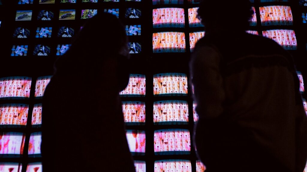 Two people silhouetted in front of a wall of glowing video screens in a dark room